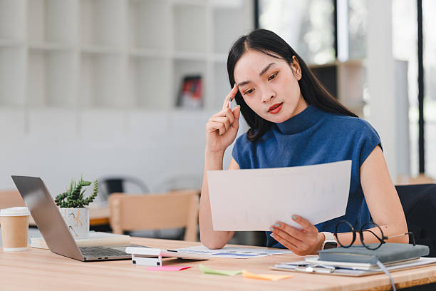 istock Asian businesswoman in blue top sits at desk, looking stressed while using calculator, surrounded by office supplies. 2221310323