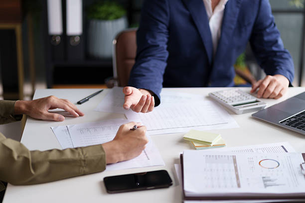 istock Two individuals engaged in a discussion at a desk, with a focus on their hands and a document. A laptop displaying charts and graphs is visible in the background. 2221317011