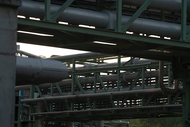 A striking view of an old, rusted industrial fabric pipe within the iconic Landschaftspark Duisburg-Nord, Germany. This former ironworks site has been transformed into a public park and cultural space, preserving the raw beauty of its massive steel structures and aged machinery. The towering chimney and intricate network of rust-covered pipes and walkways capture the essence of industrial heritage and urban decay. Set against a lush green backdrop and cloudy sky, this image embodies the contrast between nature and industry, making it ideal for themes related to history, transformation, or industrial tourism