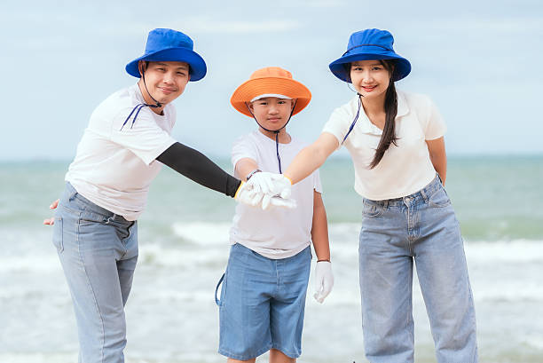 Three volunteers participate in a beach cleanup by the ocean, wearing hats and gloves while collecting trash. They pose together, showcasing teamwork and environmental awareness.