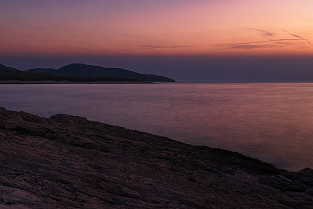 A serene, moody seascape from Hvar Island, Croatia, just moments after sunset. The sky is painted in soft purples and orange-pink hues, blending harmoniously over a calm, glassy sea. The rocky foreground leads the viewer's eye toward a darkened headland on the horizon, shrouded in shadow. This image evokes peacefulness and introspection, perfect for themes of meditation, travel, or natural beauty during golden hour.