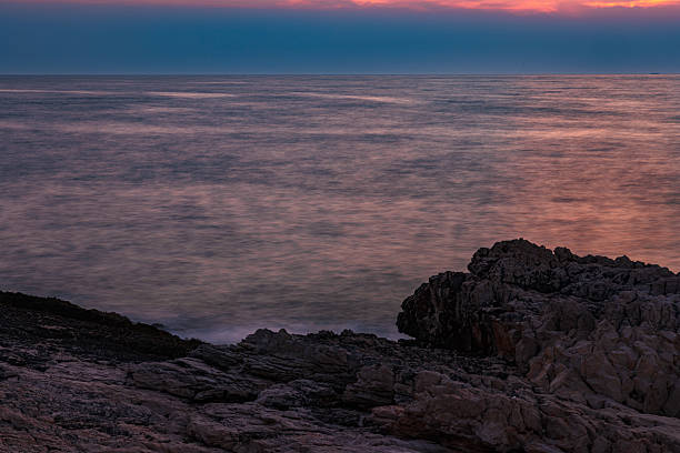 A richly textured coastal scene from Hvar Island, Croatia, captured at dusk. The warm hues of the setting sun reflect beautifully off the calm Adriatic waters, while rugged limestone formations dominate the foreground. The small stone slab near the shoreline adds depth and a hint of human touch to the natural composition. The image conveys serenity, timelessness, and the raw beauty of Mediterranean nature.