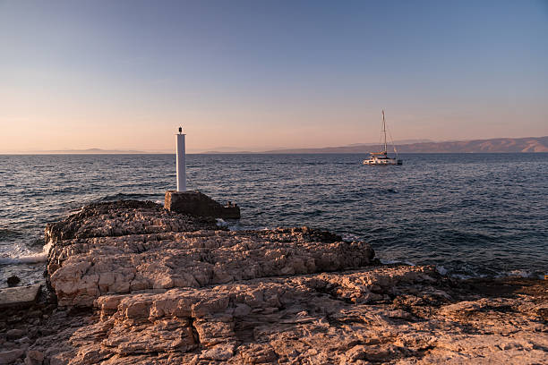 A tranquil sunset moment captured on the coast of Hvar Island, Croatia, where a small white lighthouse stands firmly on rugged natural rock formations. In the distance, a sailing catamaran drifts across the calm Adriatic Sea under a pastel sky. The golden light of summer bathes the rocks and sea, creating a warm, idyllic atmosphere. This peaceful maritime landscape blends natural textures and nautical elements, perfect for travel promotions, luxury getaway ads, or coastal-themed editorial content.