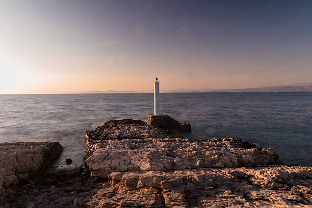 A minimalist coastal scene from Hvar Island, Croatia, showcasing a narrow white lighthouse standing solitary on a rugged outcrop of layered stone. The calm Adriatic Sea stretches endlessly under a soft sunset sky, casting warm golden light across the textured rock. The image radiates peace, isolation, and meditative calm—ideal for travel promotions, minimalist nature art, wellness themes, or ocean-inspired lifestyle editorials. The hazy outlines of distant islands add depth and dreamy atmosphere to this summer seascape.