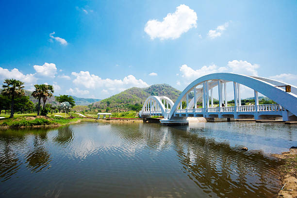 Tha Chomphu Bridge  over river Tha in Lamphun province. Curved bridge was  built in 1919 next to the Khun Tan Tunnel. Bridge crosses the Tha River and passes through the middle of an open field and landscape  scenery. Bridge is not made with steel but  it has a reinforced concrete frame of 87.3 metres long. A view people are between bridge arches taking photos. Bridge is a travel destination.  People are part of larger group which has left a train which has stopped at bridge for letting people go to lookout travel destination.