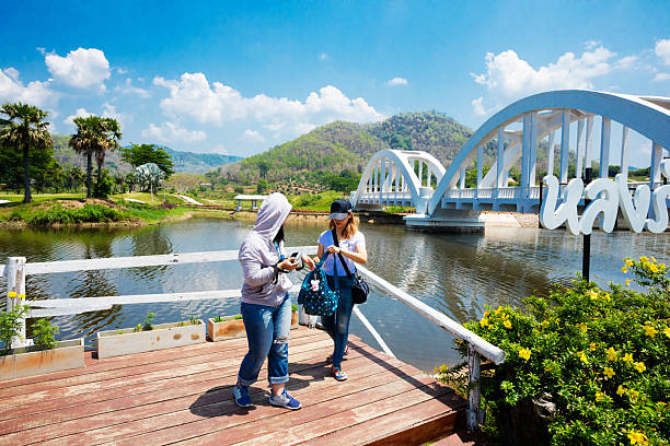 Two thai women at river Tha and Tha Chomphu Bridge  in  Lamphun province. The bridge was  built in 1919 next to the Khun Tan Tunnel. Bridge crosses the Tha River and passes through the middle of an open field and landscape  scenery. Bridge is not made with steel but  it has a reinforced concrete frame of 87.3 metres long. Women are standing with bags and camera on small wooden platform at riverside. They are taking photos with bridge and landscape. People are part of larger group which has left a train which has stopped at bridge for letting people go to lookout travel destination. OIne woman is wearing a cap