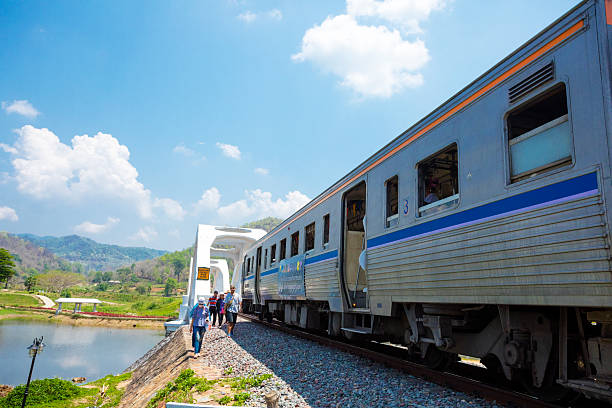 Stopped train at Tha Chomphu Bridge in Lamphun province at sunny day. Bridge was built in 1919 next to the Khun Tan Tunnel. Bridge crosses the Tha River and passes through the middle of an open field and landscape  scenery. Bridge is not made with steel but  it has a reinforced concrete frame of 87.3 metres long. A few thai people are exiting train and are going to to many other people at lookout at river Thai below trai  and bridge.