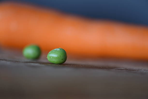 Macro photograph of a fresh green pea with water droplets on its surface, placed on a wooden background. A softly blurred orange carrot is visible in the background, creating a colorful contrast. The shallow depth of field emphasizes freshness and simplicity, suitable for healthy food and natural lifestyle themes.