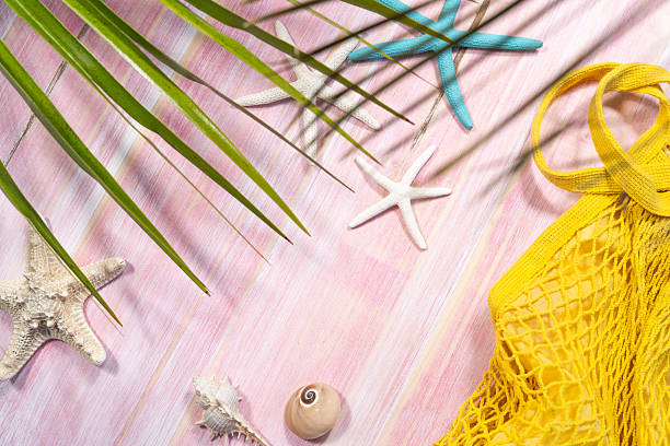 Summer beach composition featuring yellow mesh bag, seashells, starfish, palm leaf resting on pink wooden surface