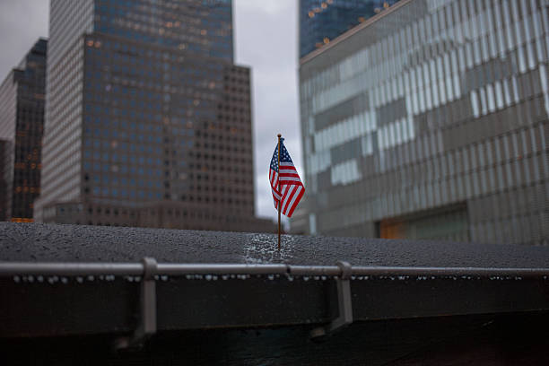 A small American flag positioned on the edge of the 9/11 Memorial in Lower Manhattan with the towering skyscrapers of the Financial District rising dramatically in the background. The flag serves as a poignant symbol of remembrance against the imposing glass and steel architecture under an overcast sky. The contrast emphasizes the memorial's solemn significance within the bustling urban environment.