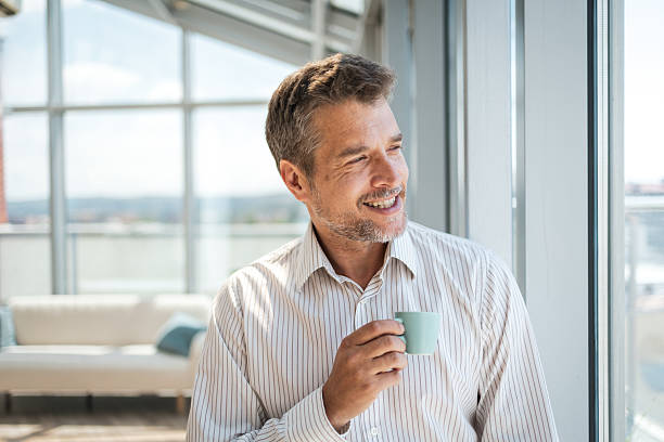 istock Man enjoying a coffee while smiling and looking outside next to a window 2221370380