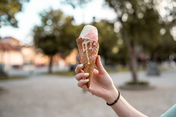 Close-up of a hand holding a melting ice cream cone with trees blurred in the background, evoking summer vibes, relaxation, and indulgence during a sunny day in a park.