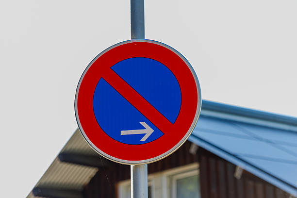 A red and blue no parking sign with a white arrow is mounted on a metal pole with a building visible in the background. Sign 63.
