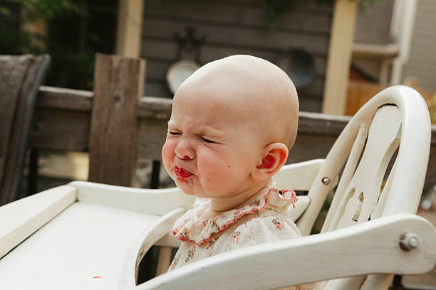 A baby girl, seven months old, makes a sour face as she has her first taste of strawberries. She is sitting in a wooden highchair on a back deck. Shot with a Canon 5D Mark IV.