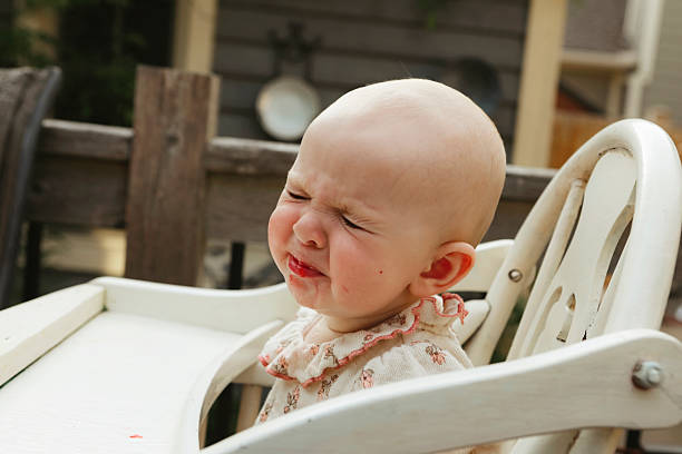 A baby girl, seven months old, makes a sour face as she has her first taste of strawberries. She is sitting in a wooden highchair on a back deck. Shot with a Canon 5D Mark IV.