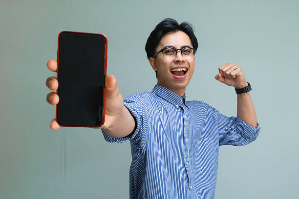 istock Happy excited young Asian man celebrating success while showing a blank smartphone screen, making a victory gesture on an isolated background, perfect for ads or promotions 2221416097