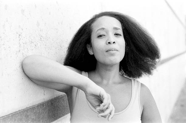 Grainy monochrome analog film photograph of a young woman with voluminous curly hair leaning against a textured wall, her serious expression and relaxed posture creating a contemplative mood