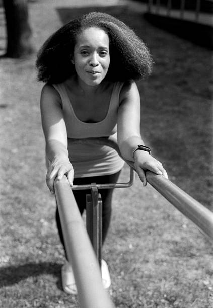 Young woman with curly hair is holding onto parallel bars, performing rehabilitation exercises outdoors, captured in a vintage, grainy black and white style