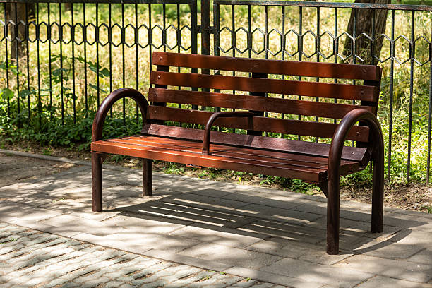 A weathered wooden bench sits empty beside a chain barrier along a paved roadway. The scene captures a bright sunny day, highlighting the urban landscape and seating area.