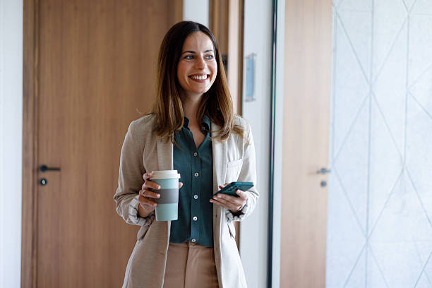 istock Young businesswoman walking through office hallway holding reusable coffee cup and using smartphone 2221468203