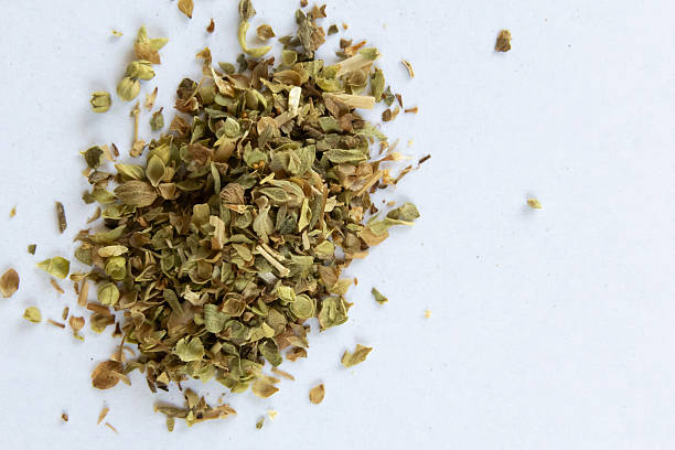 Close-up of a small pile of dried oregano leaves scattered on a clean white surface. The herb's natural green and brown tones are clearly visible, emphasizing its texture and organic quality. The minimalist composition highlights the simplicity and culinary use of dried herbs.