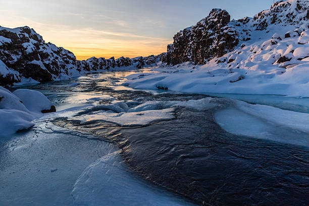 Winter outdoor scene showing a snow-covered river near the partially frozen Oxararfoss Waterfall in Thingvellir national park in the Golden circle in Iceland.