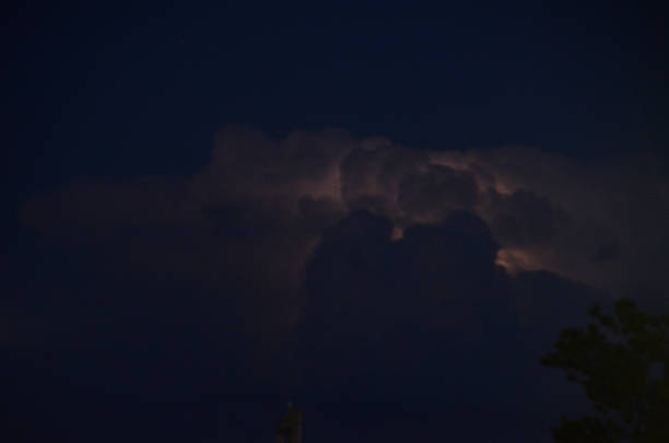 A powerful bolt of lightning forks across a dark, cloudy sky above silhouetted trees and distant hills during a nighttime storm.