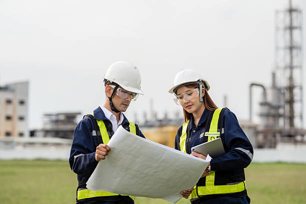 istock Engineering Team Analyzing Blueprint and Tablet Data at an Industrial Site. Two people wearing safety gear and looking at a drawing 2221489085