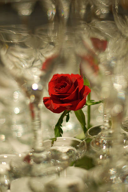 Abstract rendering of a red rose bud in a clear glass vase set among clear glass stemware stock photo