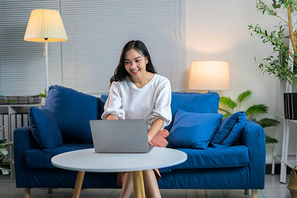 istock Young Asian woman using laptop, working remotely from home, sitting on comfortable blue couch in cozy living room 2221497791