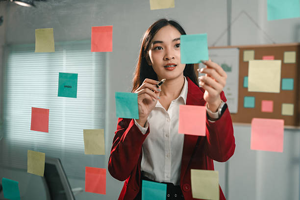 istock Young businesswoman is using colorful sticky notes on a glass wall to plan and organize tasks, showcasing a visual approach to project management in a modern office setting 2221497934
