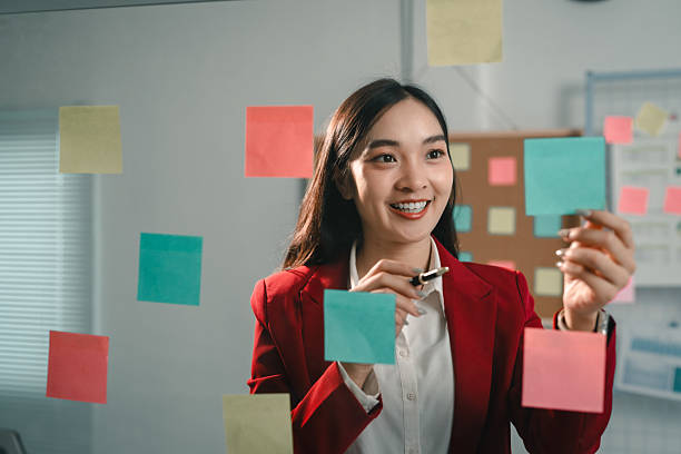 Young businesswoman is smiling while using sticky notes on a glass wall during a brainstorming session