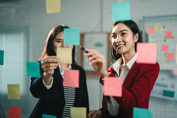 istock Two young businesswomen smiling and using colorful sticky notes on a glass wall during a creative meeting in the office 2221497957