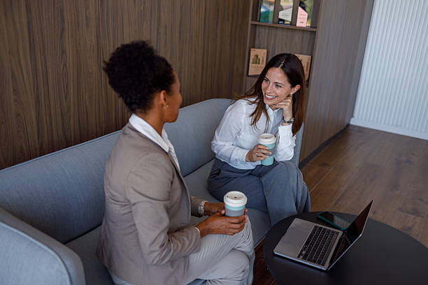 istock Professional women drinking coffee, conversing while relaxing on sleek lobby sofa during workplace break 2221505640