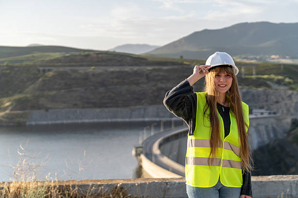 istock Smiling young blonde female engineer adjusting her hardhat at a dam construction site, with a clear sky, water, and mountain landscape in the background 2221507571
