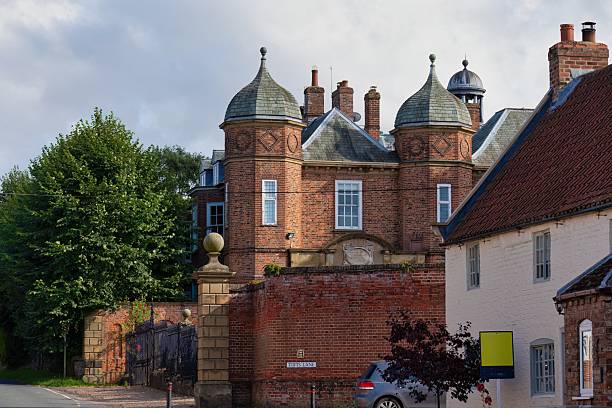 A red brick building with distinctive turrets stands among trees and other buildings under a cloudy sky in Brafferton - North Yorkshire - United Kingdom