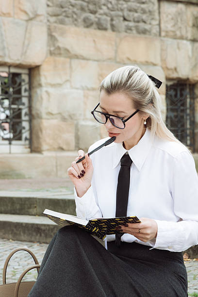 istock Professional woman in white shirt and black skirt, sitting outdoors, reading notebook while holding pen, deep in thought, preparing for summer vacation during lunch break with a serene atmosphere 2221514057