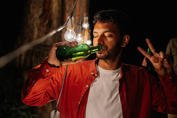 istock Young man drinking beer from bottle at night and making peace sign 2221541706