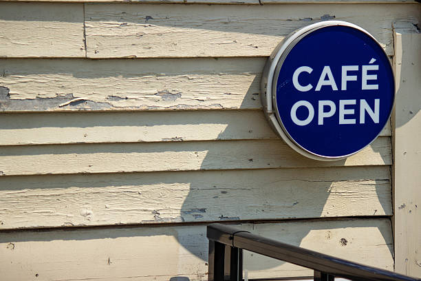 A blue circular “Café Open” sign mounted on a weathered wooden wall, indicating that a café is open for business. Taken in natural daylight, this image conveys a welcoming, local, and rustic feel.