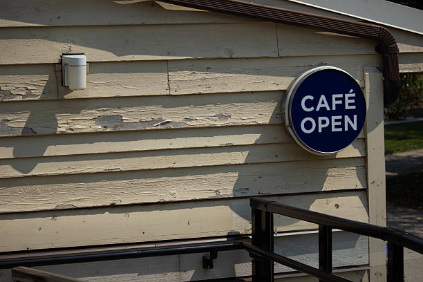 A blue circular “Café Open” sign mounted on a weathered wooden wall, indicating that a café is open for business. Taken in natural daylight, this image conveys a welcoming, local, and rustic feel.
