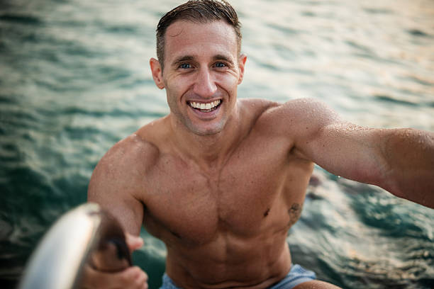 Shirtless tourist taking a selfie coming out of the water, holding the selfie stick with one hand and the ladder with the other
