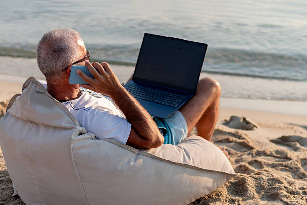 istock Mature man with laptop talking by mobile phone on beach. 2221559551