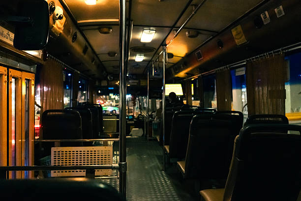 A quiet, empty public city bus in Bangkok at night. The image captures rows of vacant seats, soft yellow lighting, and a calm city ambiance visible through the windows.