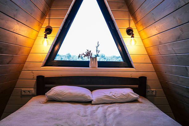 istock Warm bedroom interior in wooden A-frame cabin, bed with pillows, triangular window with forest view, cozy lighting with wall lamps, natural wood decor 2221583793