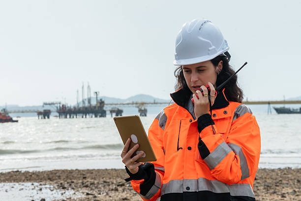 istock Female engineer using a tablet and walkie-talkie for a field report at a maritime construction site. Woman technician multitasking with mobile technology for data entry and communication offshore. 2221584264
