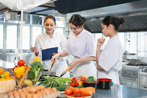 istock Young Chefs Learning to Cook with Fresh Ingredients, Culinary Instructor Guides Students in a Professional Kitchen, Women in Chef Uniforms Preparing Food in a Commercial Kitchen 2221584443