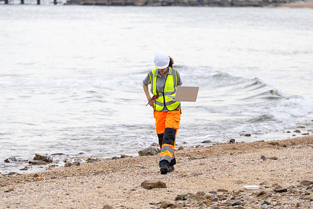 istock Environmental Scientist Conducting Beach Cleanup Survey, Female Engineer Using Laptop for Coastal Area Assessment, Worker in Safety Vest and Hard Hat Documenting Outdoor Site 2221594500