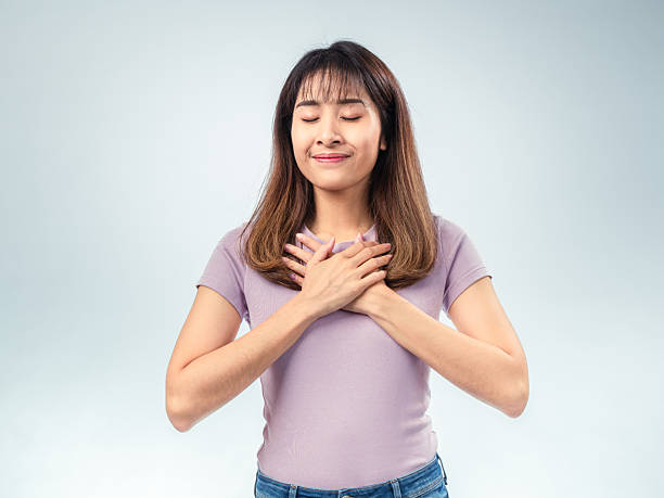 istock Asian woman smiling with both hands placed over her chest, expressing gratitude or appreciation while wearing a purple t-shirt and jeans against a light studio background 2221596003