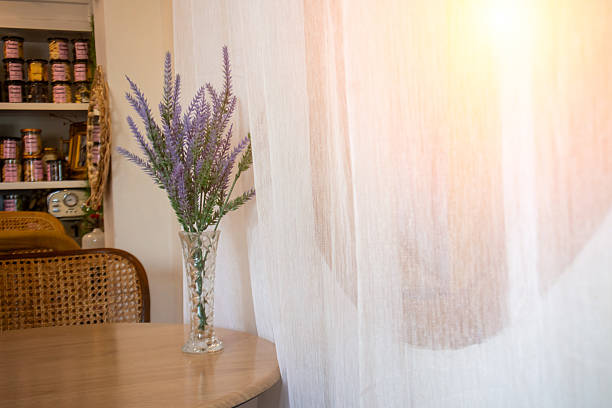 A beautiful lavender bouquet in a clear vase sits on a wooden table by a sheer curtain, creating a calm and inviting atmosphere in a bright interior space.