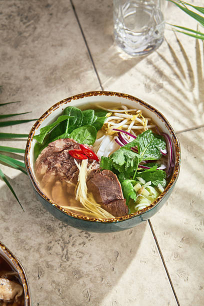 Pho bo soup in sunlight with rustic overhead composition, showcasing herbs, beef, and vibrant garnishes